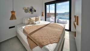 A modern bedroom with a large bed, a view of the sea through a sliding door, and decorative wall art. - Villa Saturday in Foinikas (Agios Vasileios)