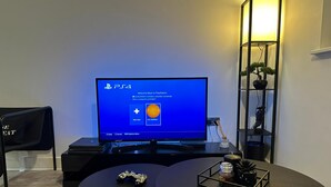 En PlayStation 4-konsol på en tv-skärm, en spelstol, ett runt bord med en spelkontroll och en liten växt, och en hög, svart hylla med dekorativa föremål.