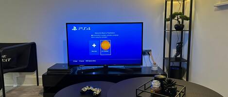Una consola PlayStation 4 en una pantalla de televisor, una silla gamer, una mesa redonda con un control de juego y una planta pequeña, y un estante negro alto con objetos decorativos.