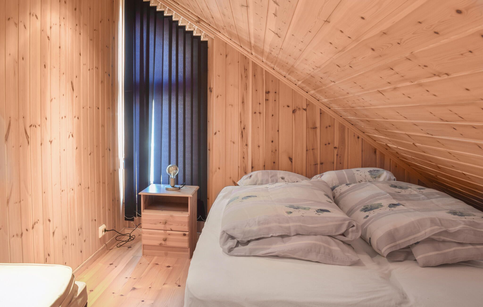Un dormitorio con techo inclinado, paredes de madera, una cama con ropa de cama floral, una mesita de noche con lámpara y una ventana con persianas verticales.