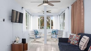 A modern living room with a dark blue sofa, two white armchairs, a ceiling fan, a flat-screen TV, and a large window with curtains. - Zana del Mar | Steps from the Beach (Humacao)