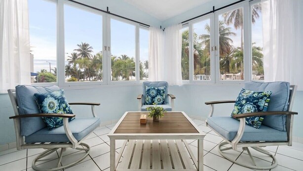 A room with two rocking chairs, a wooden table, and large windows offering a view of palm trees and the sea. - Zana del Mar | Steps from the Beach (Humacao)