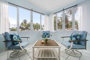 Una habitación con dos mecedoras, una mesa de madera y ventanas amplias que dan a palmeras y al mar.