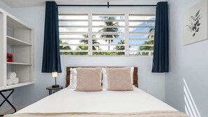 A hotel room with a bed, a nightstand, a shelf with towels, and a window with curtains. - Zana del Mar | Steps from the Beach (Humacao)