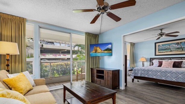 Habitación de hotel con piso de madera, un sofá con cojines amarillos, una mesa de centro de madera, un televisor en la pared, una cama con colcha estampada y un balcón con vistas a palmeras.