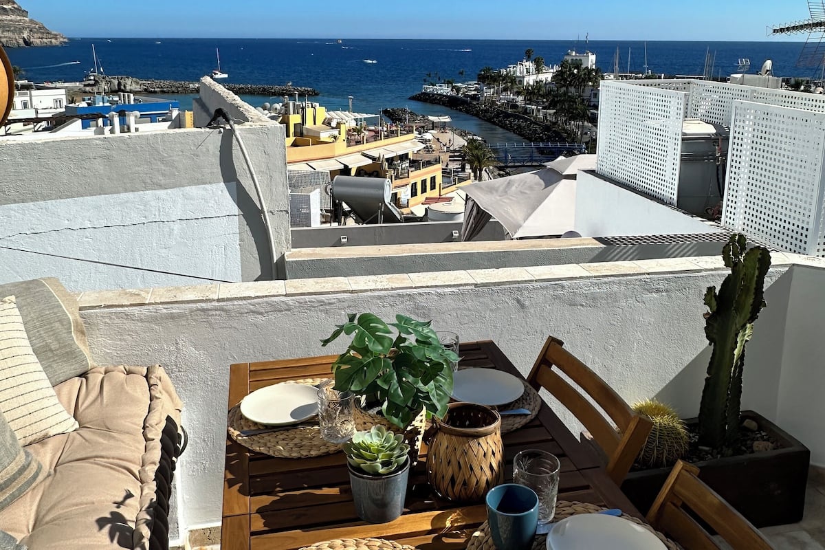 A dining table set with plates, glasses, and a potted plant, overlooking a coastal view with buildings and the sea.