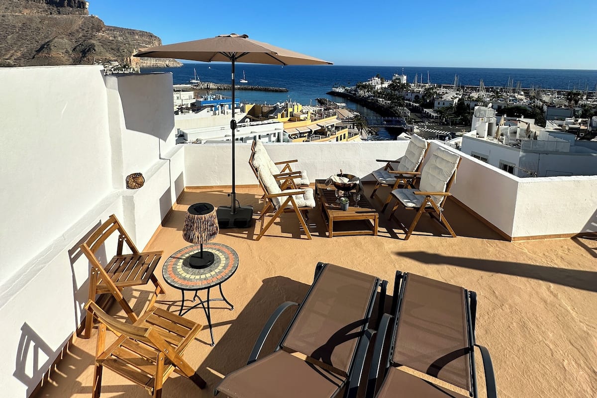 A rooftop terrace with sun loungers, a table, and a view of the sea and buildings.
