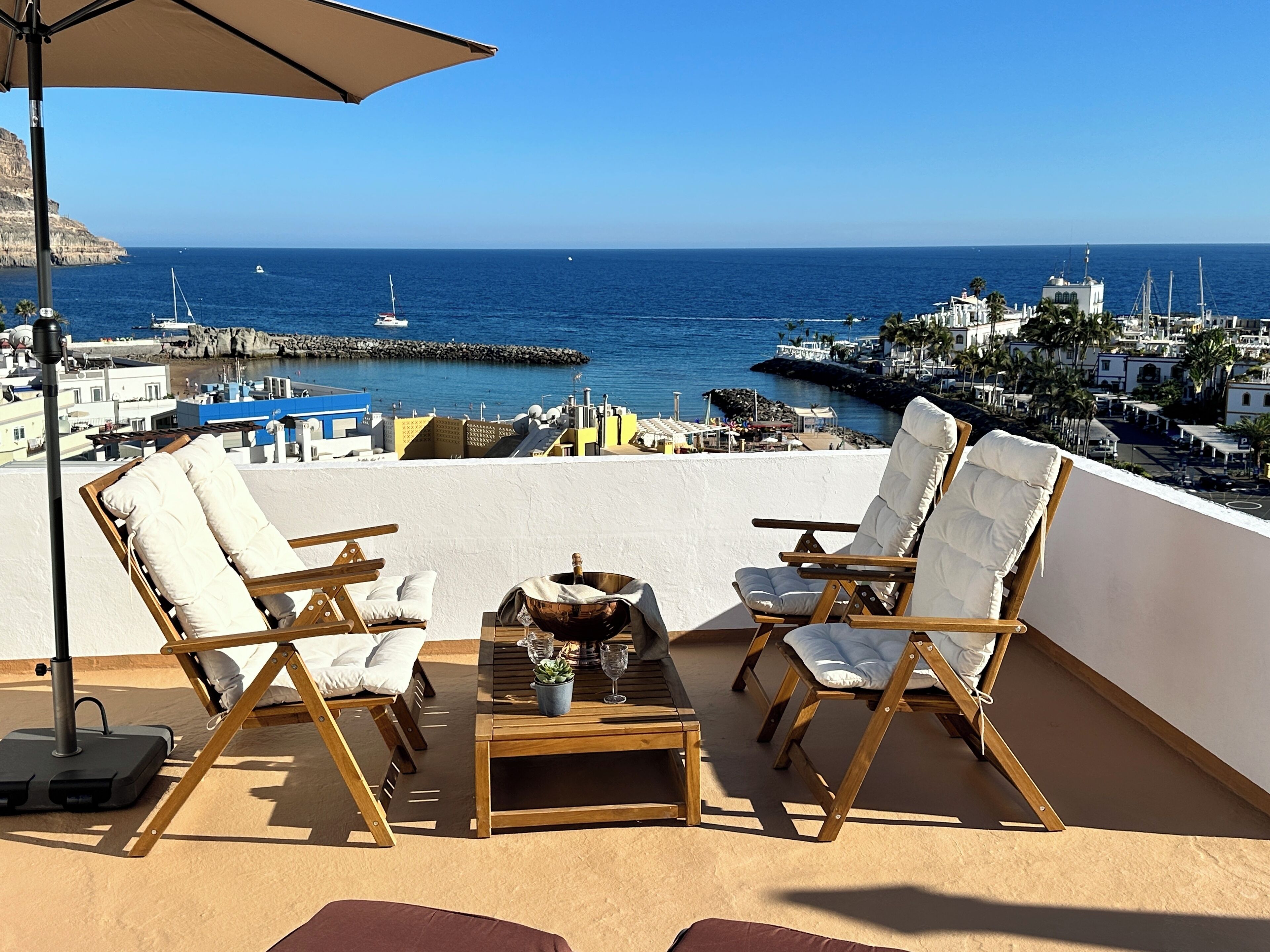 A terrace with two wooden lounge chairs, a table with a bottle and glasses, and a view of the sea and buildings.