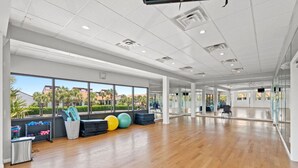 A spacious gym with wooden floors, exercise equipment, and large windows offering a view of palm trees and buildings. - Edgewater Golf Villas 1714 | Family-Friendly Community, Beach Access, Lagoon-Style Pool (Panama City Beach)
