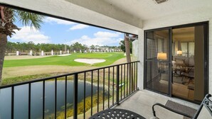 A balcony with a view of a golf course, a pool, and a dining area with chairs and a table. - Edgewater Golf Villas 1714 | Family-Friendly Community, Beach Access, Lagoon-Style Pool (Panama City Beach)
