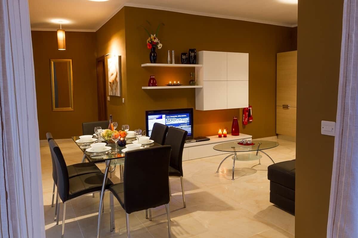 A modern dining area with a glass table, black chairs, and a TV. There are shelves with decorative items and a cabinet.