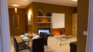 A modern dining area with a glass table, black chairs, and a TV. There are shelves with decorative items and a cabinet.