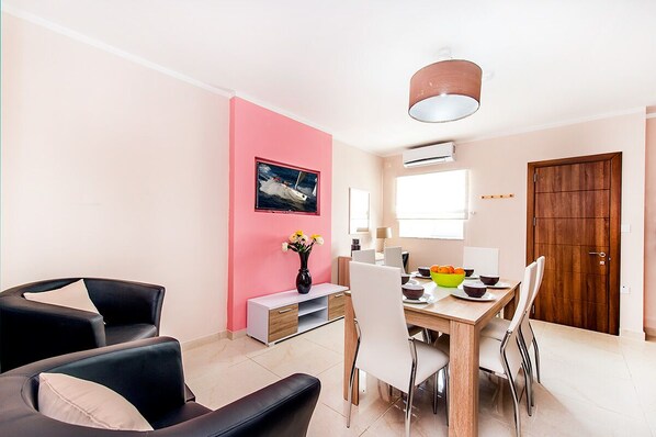 A modern living room with a pink accent wall, a dining area with a wooden table and chairs, and a large window providing natural light.
