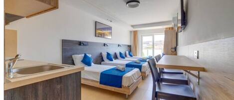 A hotel room with a kitchenette, three beds, a dining table, and a TV mounted on the wall.