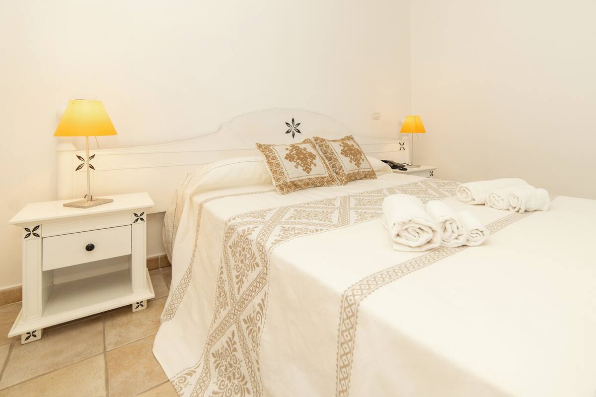 A neatly made bed with a patterned bedspread, two bedside lamps, a nightstand with a drawer, and a decorative pillow.