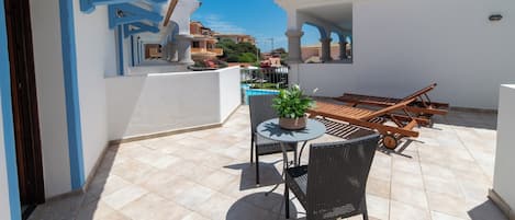 A patio with a round table, two chairs, a potted plant, and a wooden lounge chair.