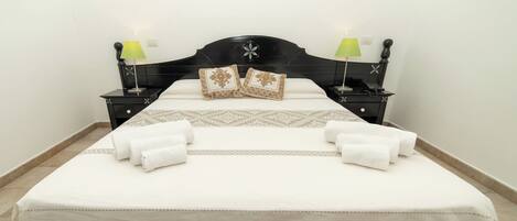 Una cama bien hecha con sábanas blancas y almohadas decorativas, dos mesitas de noche con lámparas, y una distribución sencilla y limpia del cuarto.