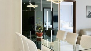 Quarto de hotel moderno com mesa de jantar de vidro, cadeiras brancas, um vaso de planta e uma grande janela com vista para a cidade.