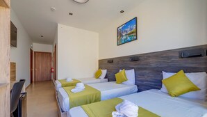 Une chambre d’hôtel avec trois lits simples, chacun recouvert d’une couverture verte et doté de draps blancs, et un chevet en bois. Un tableau représentant un paysage de montagne est accroché au mur, et un bureau en bois avec une chaise se trouve à gauche.