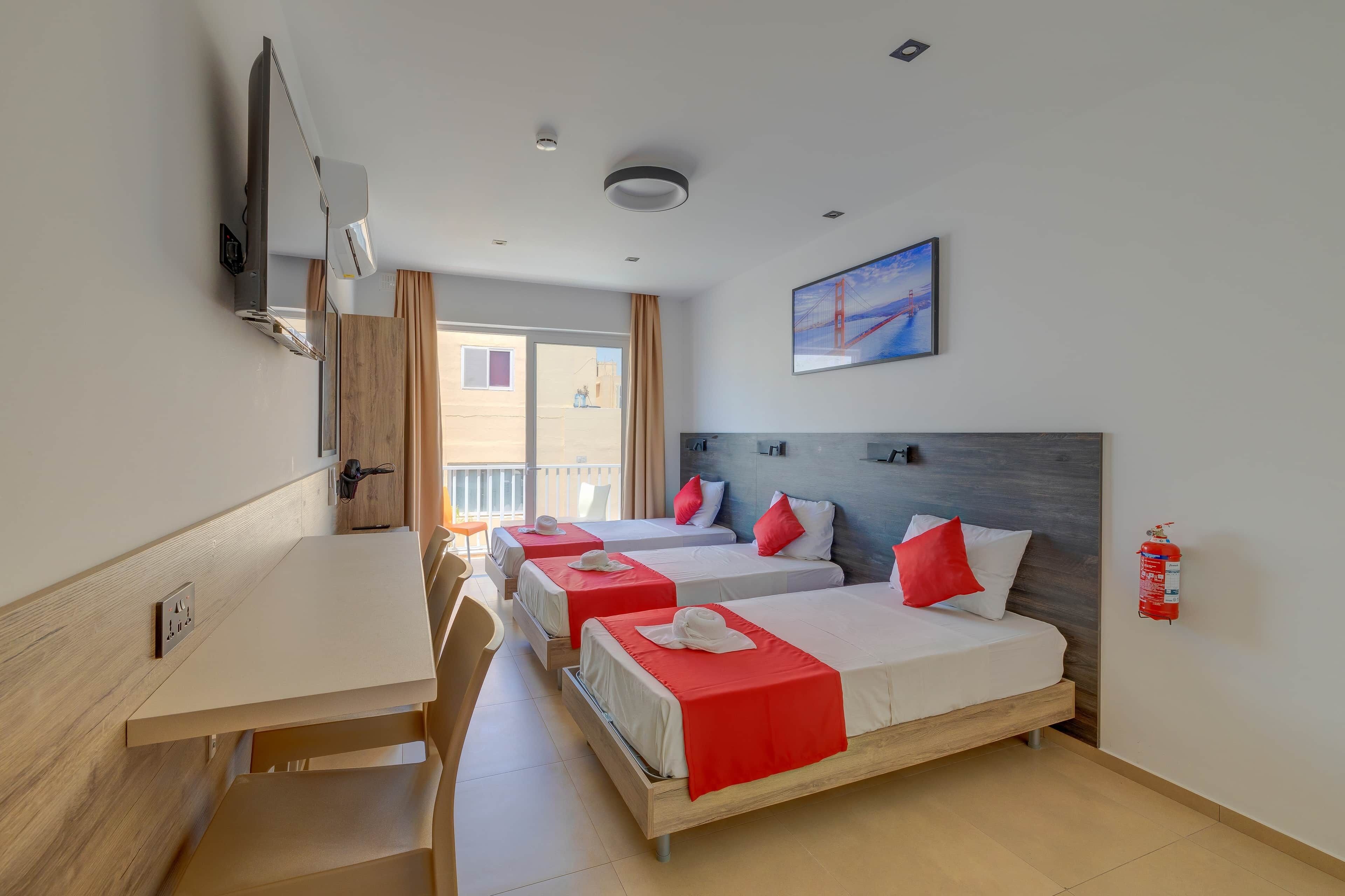 Une chambre d’hôtel avec trois lits, un bureau, une chaise, une télévision et un extincteur.