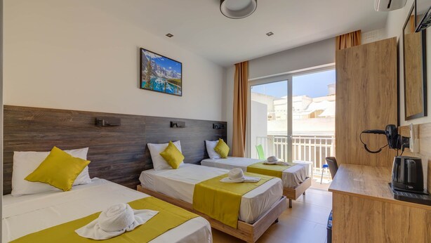 Een hotelkamer met drie bedden, een houten hoofdeinde, een schilderij aan de muur, een balkon met uitzicht en een kitchenette.