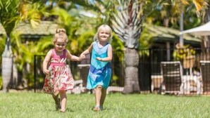 Due bambini che si tengono per mano in una zona erbosa all'aperto, con una recinzione e palme sullo sfondo.