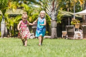 Dos niños tomados de la mano en un espacio exterior con césped, una cerca y palmeras al fondo.