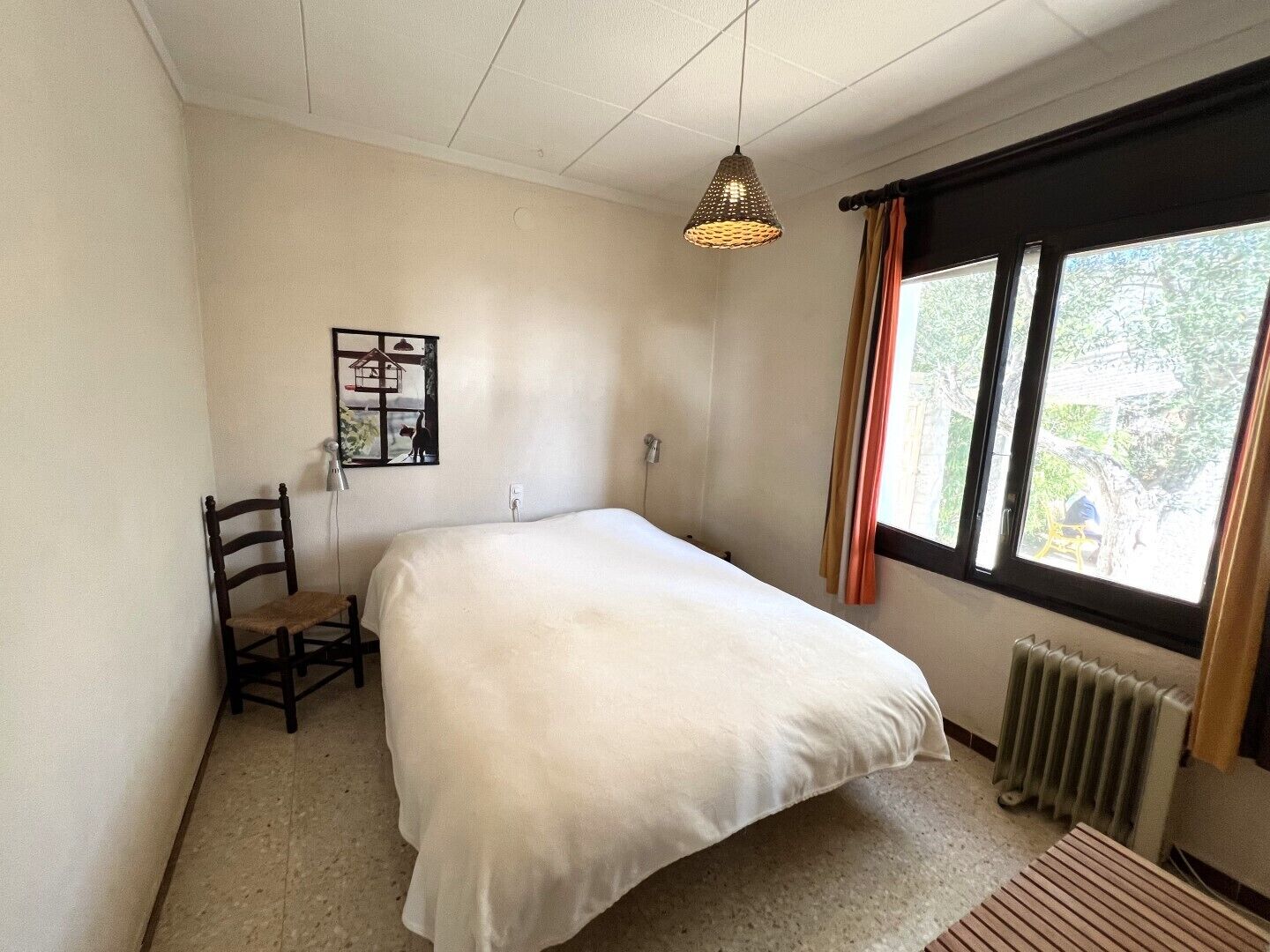 A bedroom with a bed, a chair, a radiator, a window with curtains, and a hanging light fixture.