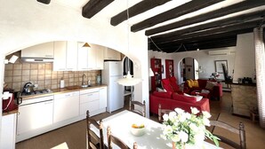 A kitchen with white cabinets, a stove, and a sink. There is a dining table with chairs and a vase of flowers. A red sofa and a red armchair are in the living area. - Rustic holiday home for 6 people, sea view, 3 bedrooms, air conditioning, Wi-Fi (El Mas Fumats)