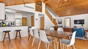A modern kitchen with white cabinets, a dining area with a wooden table and white chairs, a living room with a blue sofa, and a staircase leading to an upper level. - Lakefront 3-BR Retreat in the Berkshires (Becket)
