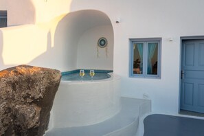 A white building with a stone structure, a circular window, and a blue door. - Sun Angelos Oia - Luxury Cave Suites (Santorini)