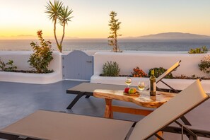 A terrace with a table set for two, a bottle of wine, and a bowl of fruit. - Sun Angelos Oia - Luxury Cave Suites (Santorini)