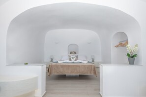 Habitación de hotel minimalista con una cama centrada bajo un techo blanco en arco, una mesita de noche pequeña, una planta en maceta y un estante con ropa fijado a la pared.