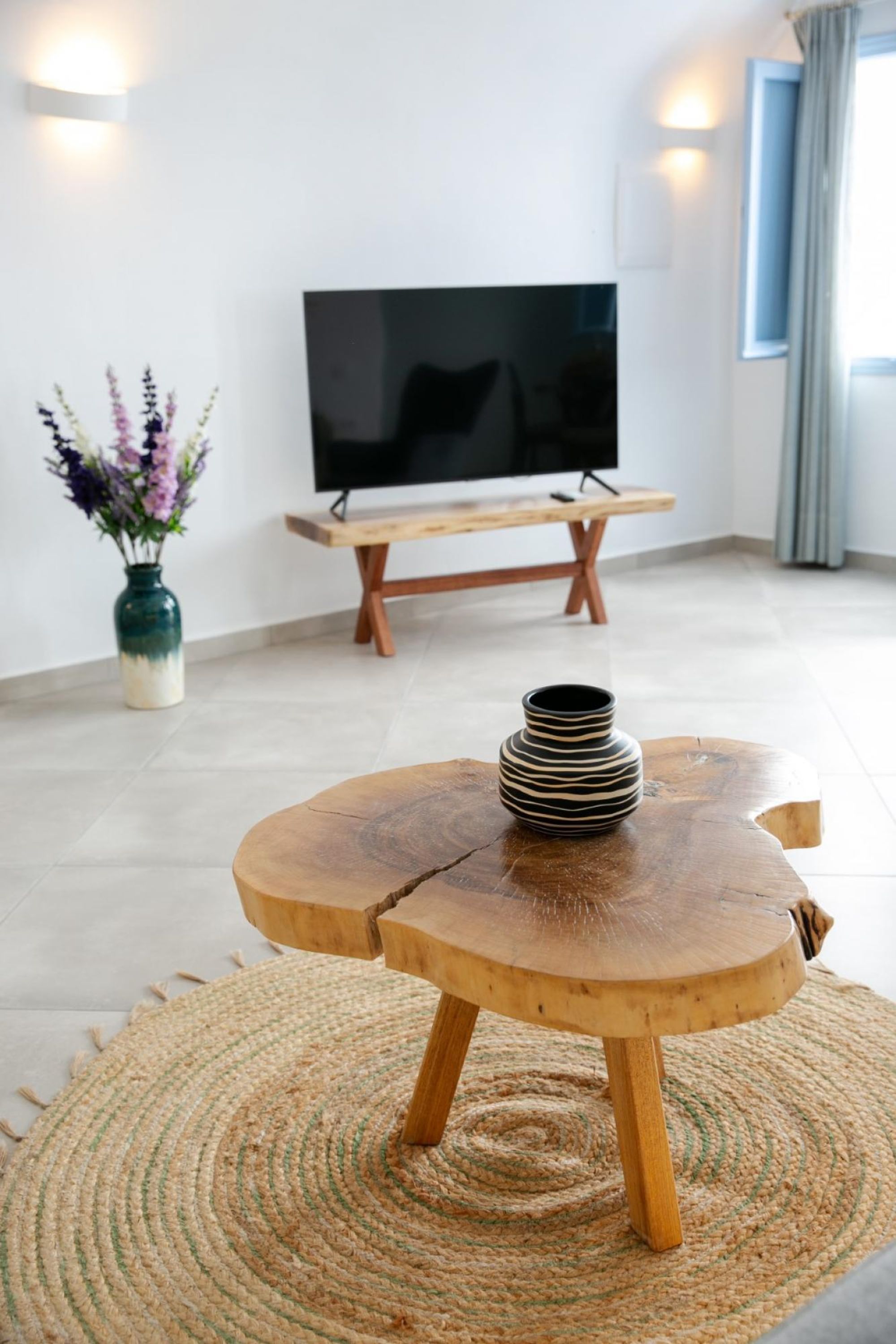 A modern living room with a flat-screen TV on a wooden stand, a round wooden table with a vase, and a woven rug.
