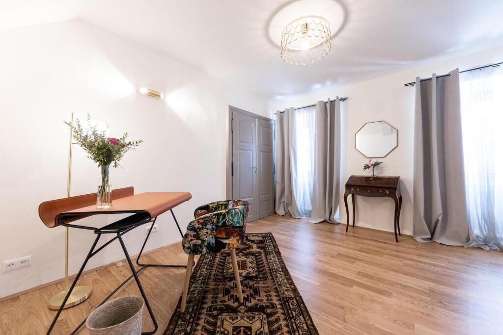 Une pièce avec un plancher en bois, une petite table sur laquelle est posé un vase de fleurs, une chaise, un tapis à motifs, un miroir sur un support en bois et une porte avec des rideaux.