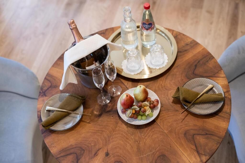 A round wooden table with a bottle of champagne, two glasses, a bottle of water, and a plate with fruits and a napkin.