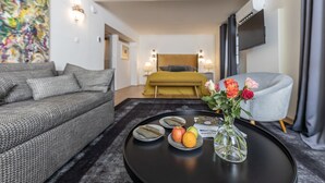 Ein modernes Hotelzimmer mit einer Couch, einem Bett mit Kopfteil, einem Esstisch mit Obst und einer Blumenvase sowie einem an der Wand befestigten Fernseher.