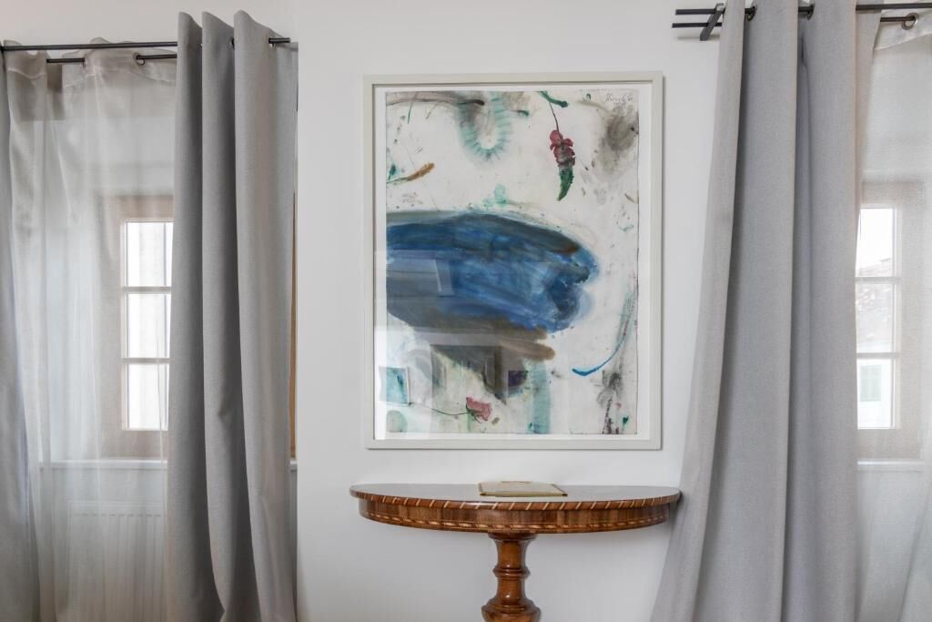Un cuadro abstracto enmarcado en una pared, una mesa redonda de madera con un libro, y cortinas de encaje en las ventanas.