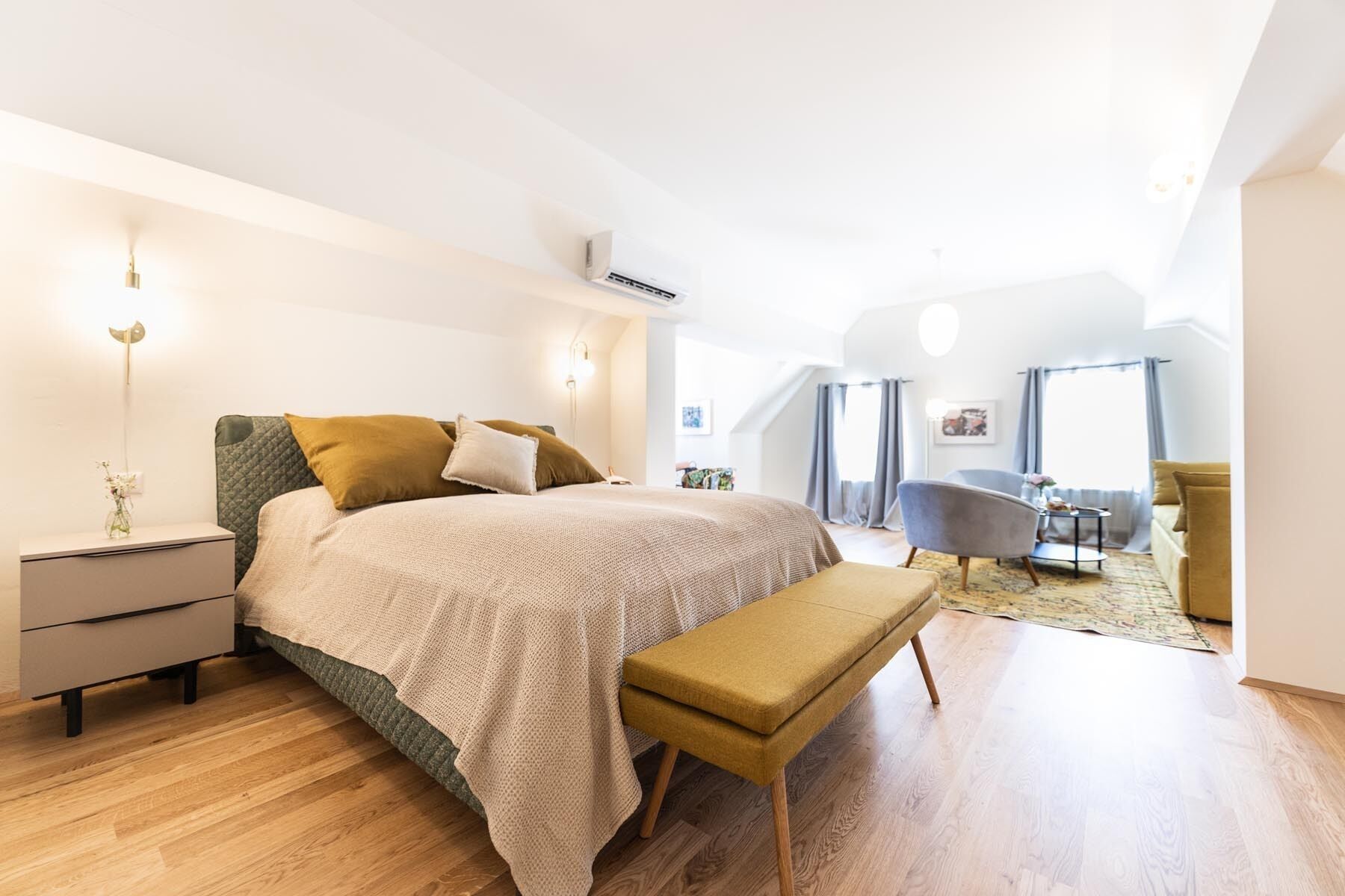 A modern bedroom with a large bed, bedside tables, a bench, and a view of a living area with a sofa and a chair.