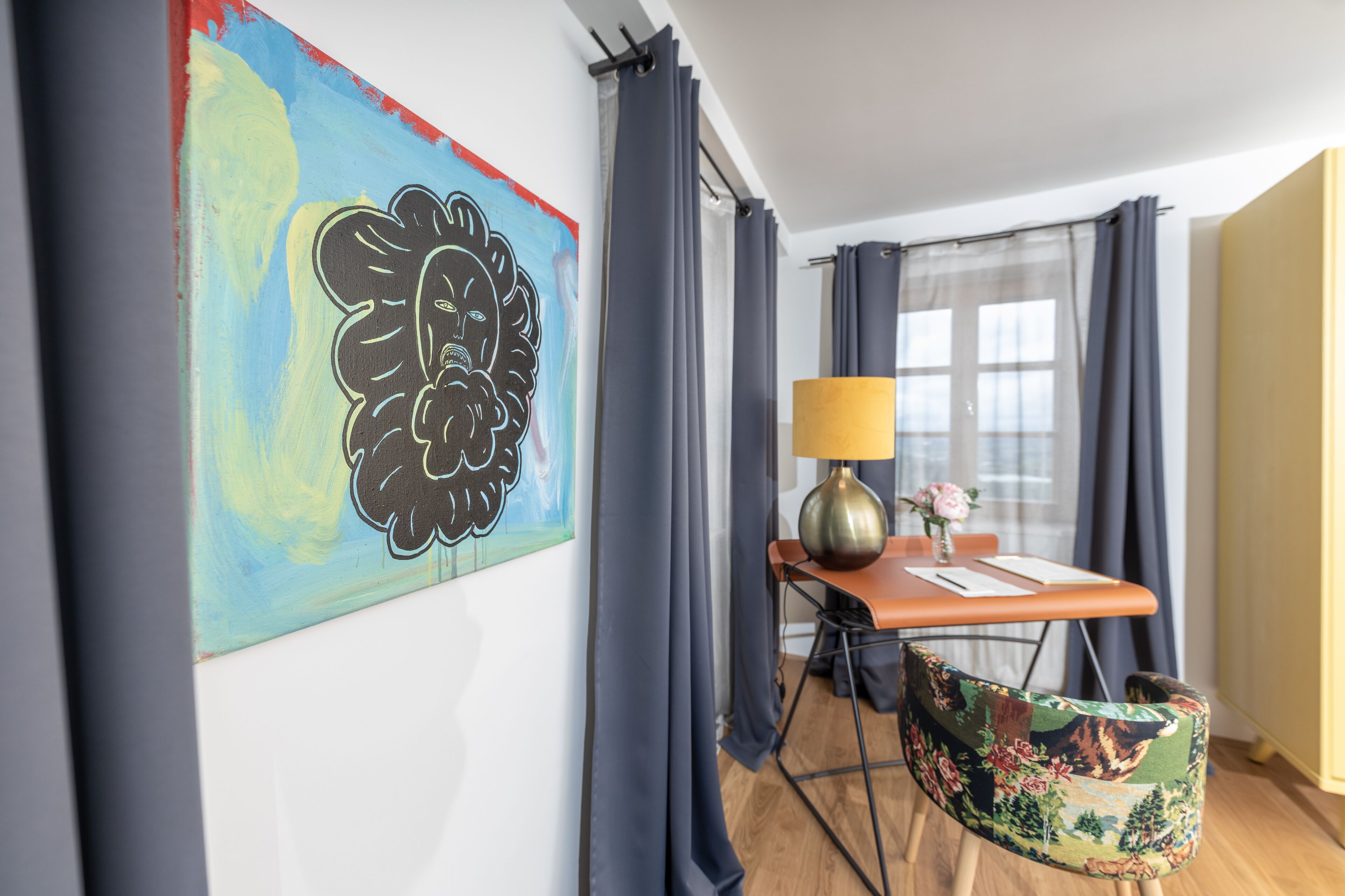 Ein Zimmer mit einem Bild an der Wand, einem Tisch mit Lampe, einem Stuhl und einem Fenster mit Vorhängen.