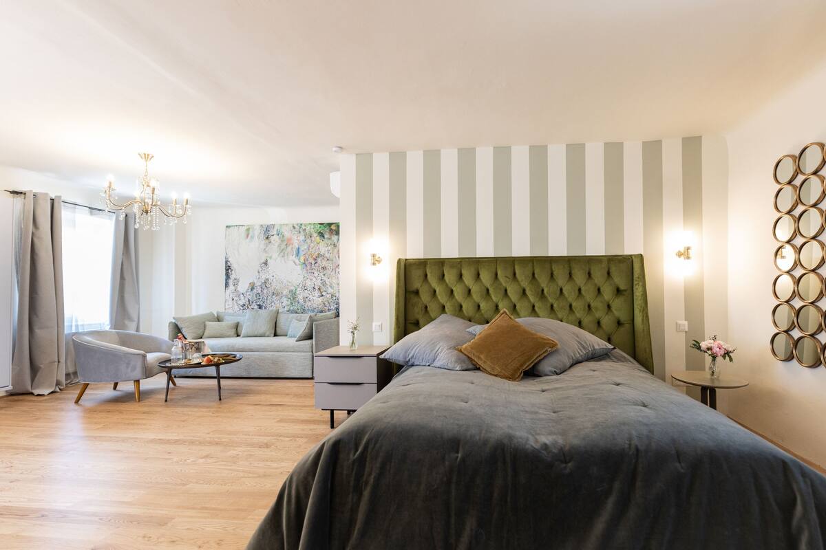 A bedroom with a tufted headboard, a grey sofa, a round coffee table, and a painting on the wall.