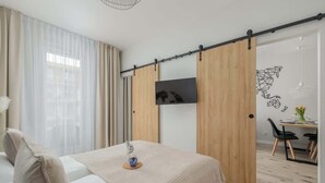 A bedroom with a bed, a TV mounted on a sliding wooden panel, a dining area with a table and chairs, and a wall with a geometric design.