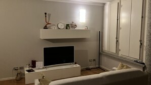 Un dormitori amb un llit, una televisió sobre un suport, una estant amb objectes decoratius i un armari.