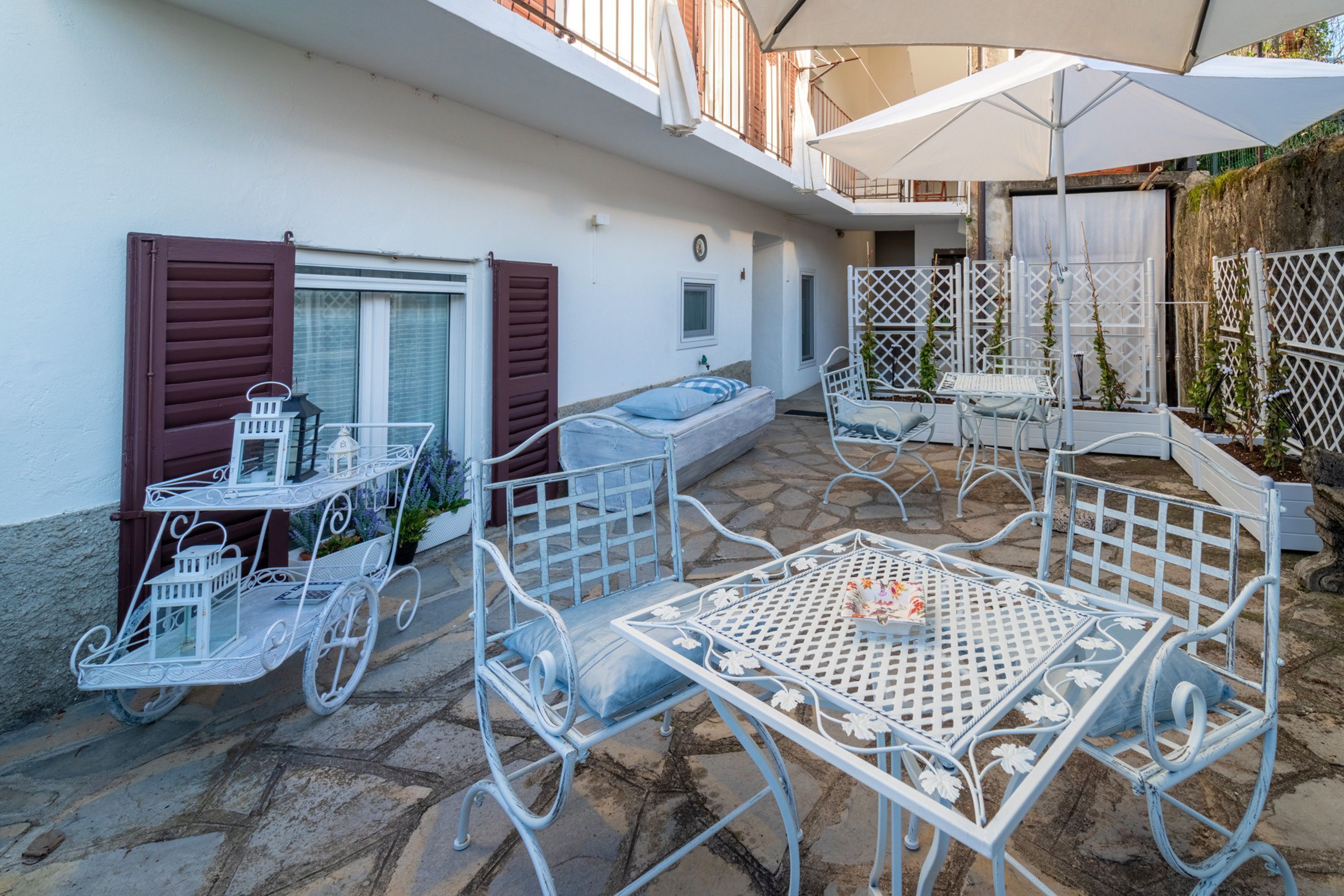 Une terrasse extérieure avec des meubles en métal blanc, notamment une table et des chaises, ainsi qu’un chariot en métal contenant des objets décoratifs.