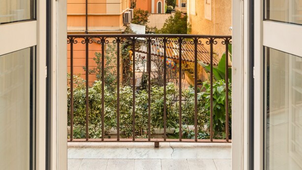 Blick von einem Balkon mit Metallgeländer auf einen Innenhof mit Gebäuden und Grünflächen.