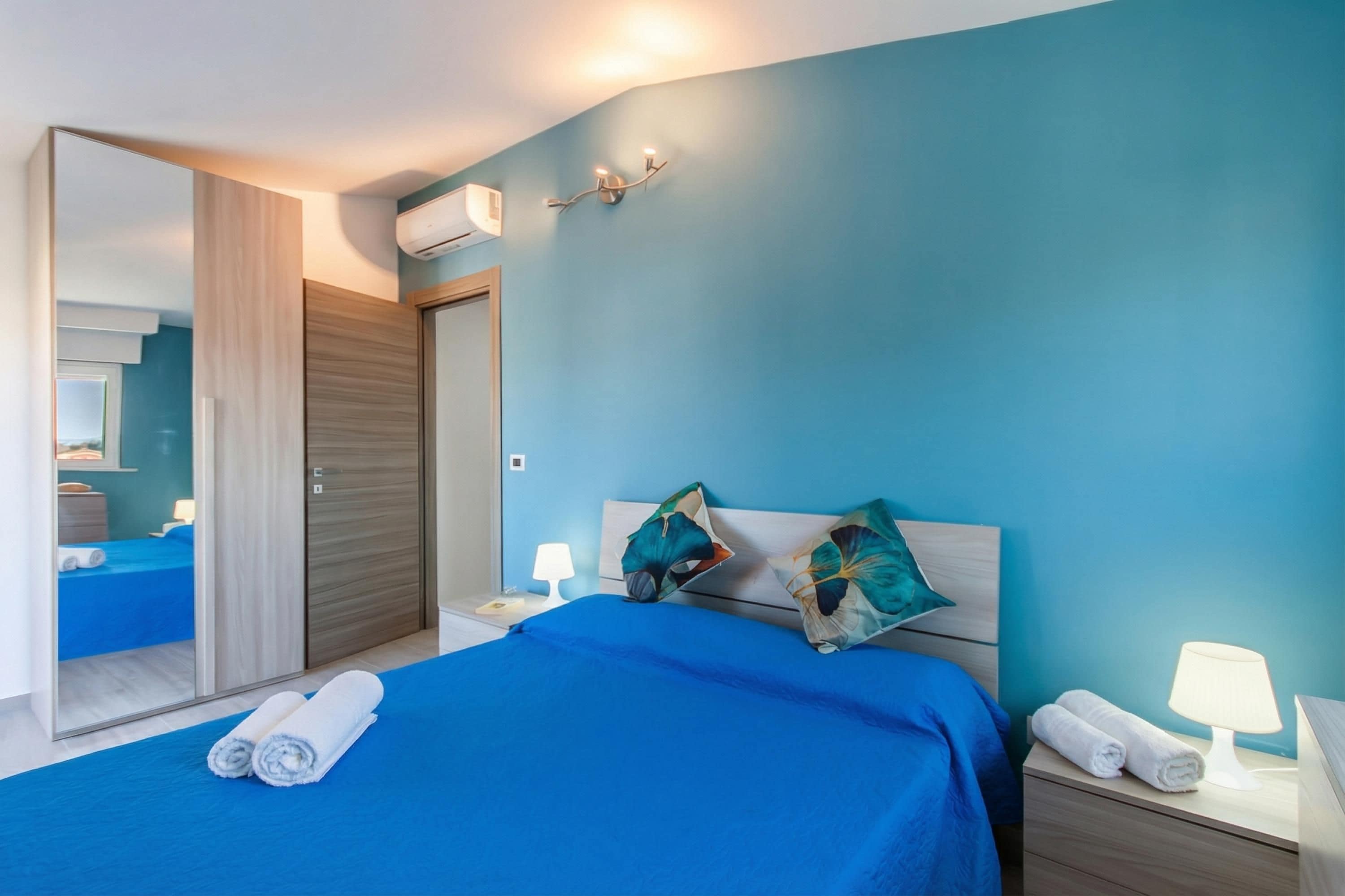 Un dormitorio con una colcha azul, dos almohadas, una mesita de noche con una lámpara, un espejo y un armario.