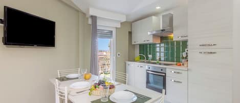 A modern kitchen with white cabinetry, a flat-screen TV mounted on the wall, and a dining area with a table set for two.