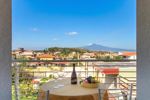 Un balcon avec une table, un verre à vin, une bouteille et un panier de fruits, d’où l’on peut admirer une ville et une montagne.