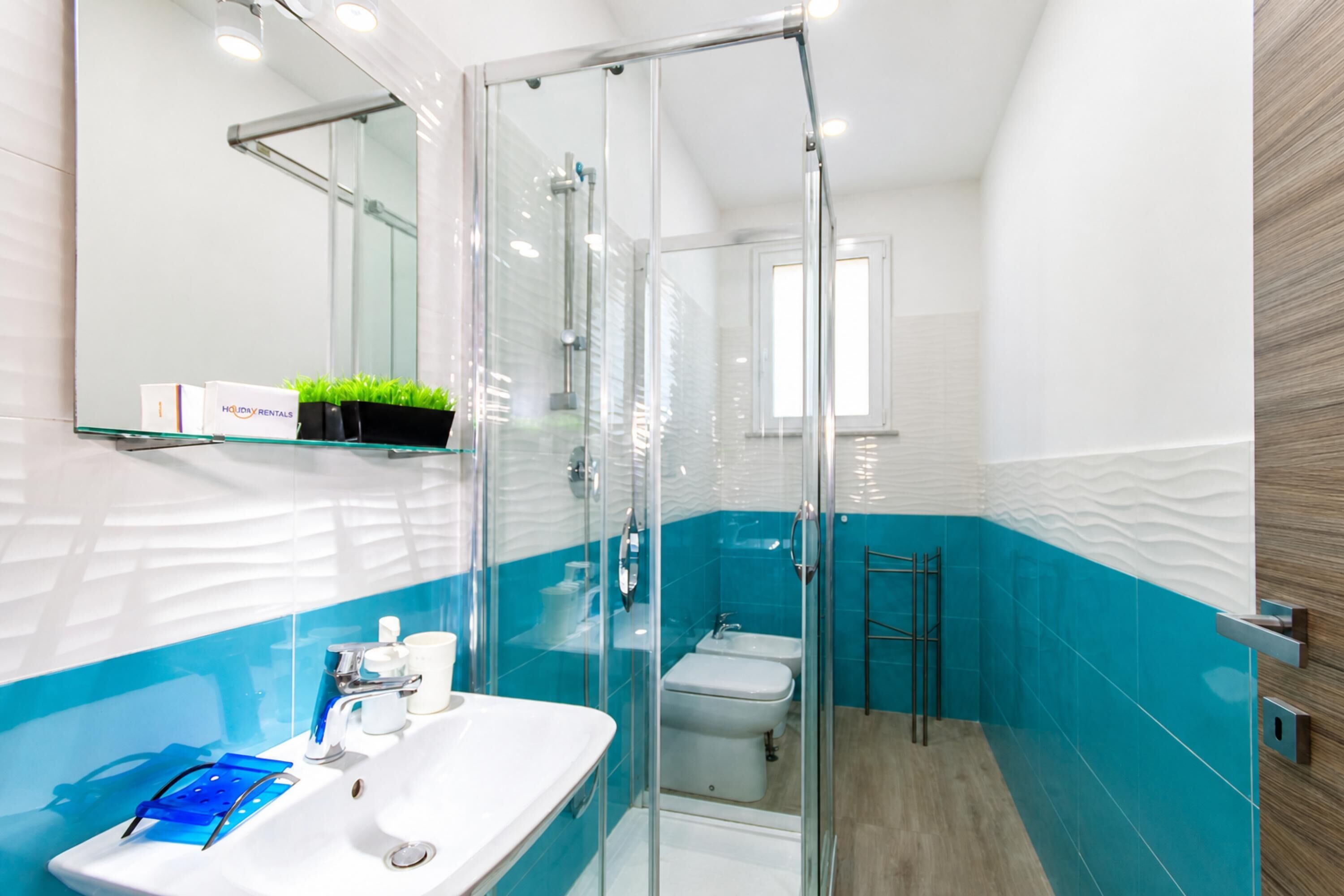 Un baño moderno con mampara de vidrio, lavamanos blanco y pared con azulejos azules.