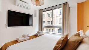 A hotel room with a bed, a flat-screen TV, a window with curtains, and a view of buildings outside. - A Breathtaking 3-BR/3BA in Trocadéro - Tour Eiffel (Paris)